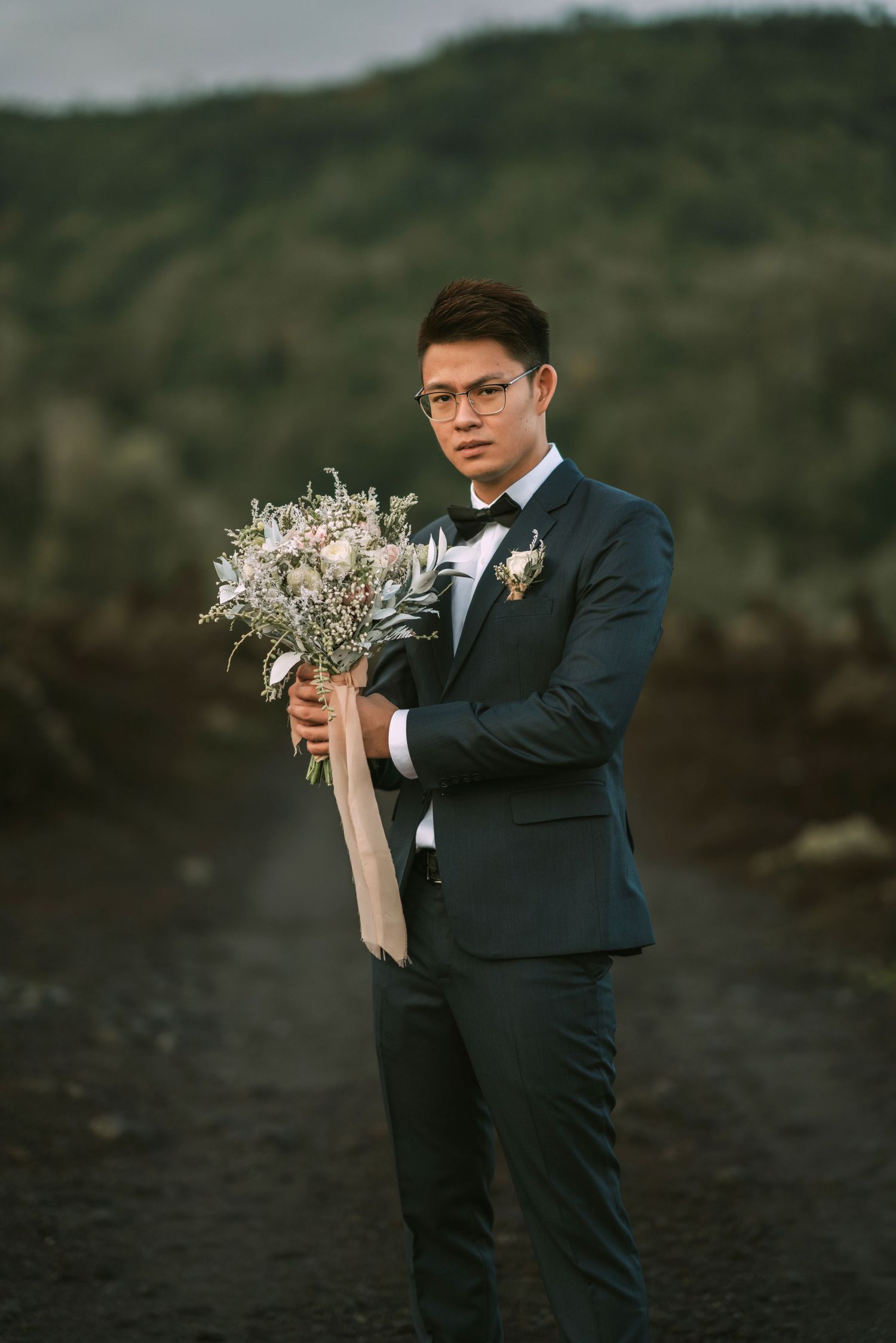 Person in dark suit holds delicate white floral bouquet against mountainous backdrop.