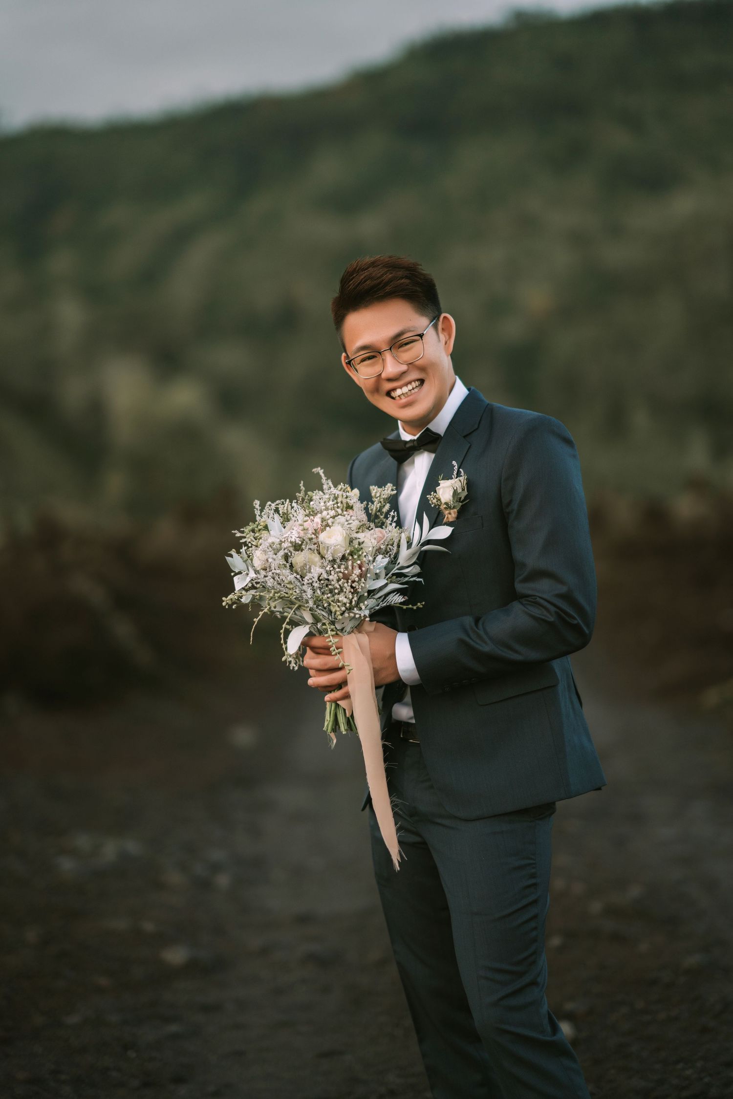 Person smiles while holding wedding bouquet on volcanic landscape with mountains in distance.