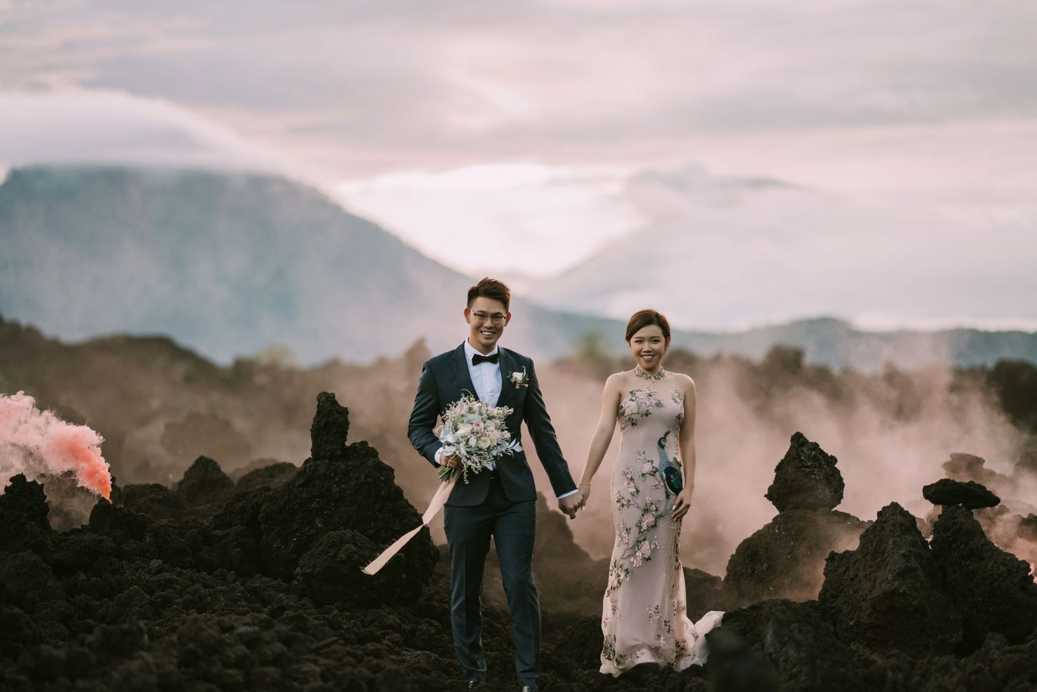 Wedding couple poses on volcanic rocks with pink sunset sky and mountain silhouettes.
