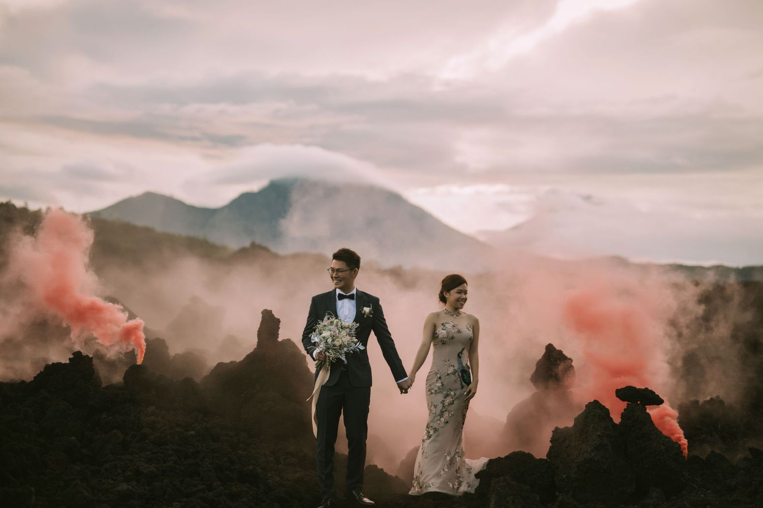 Couple holds hands on volcanic terrain with pink smoke and mountain vistas.