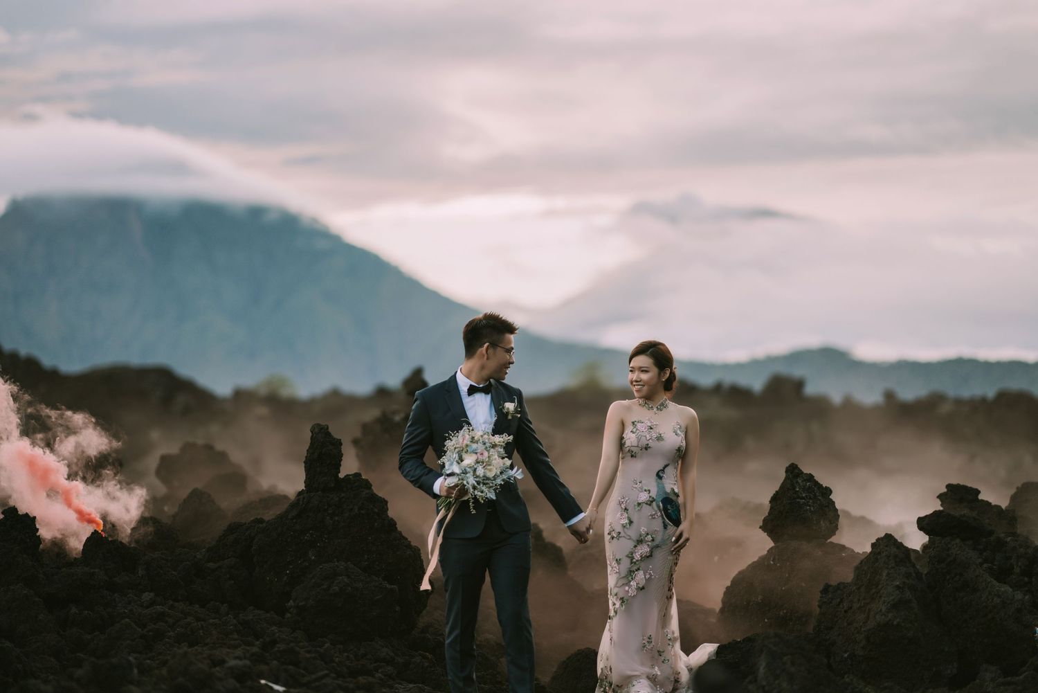 Wedding couple walks hand in hand across rugged volcanic terrain under cloudy skies.