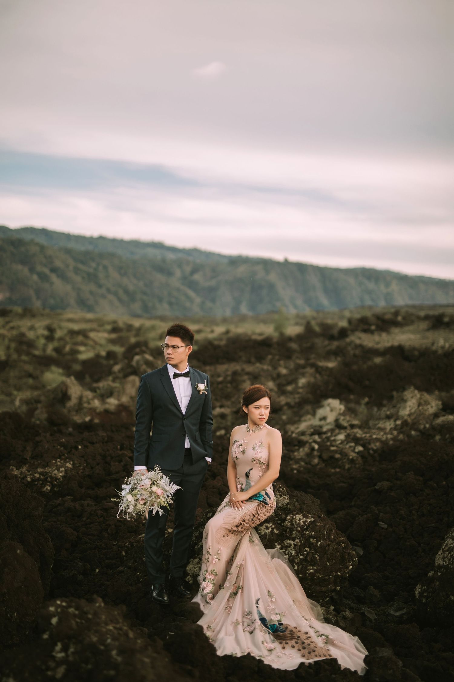 A couple poses dramatically on dark volcanic rocks with mountains in the background at sunset.
