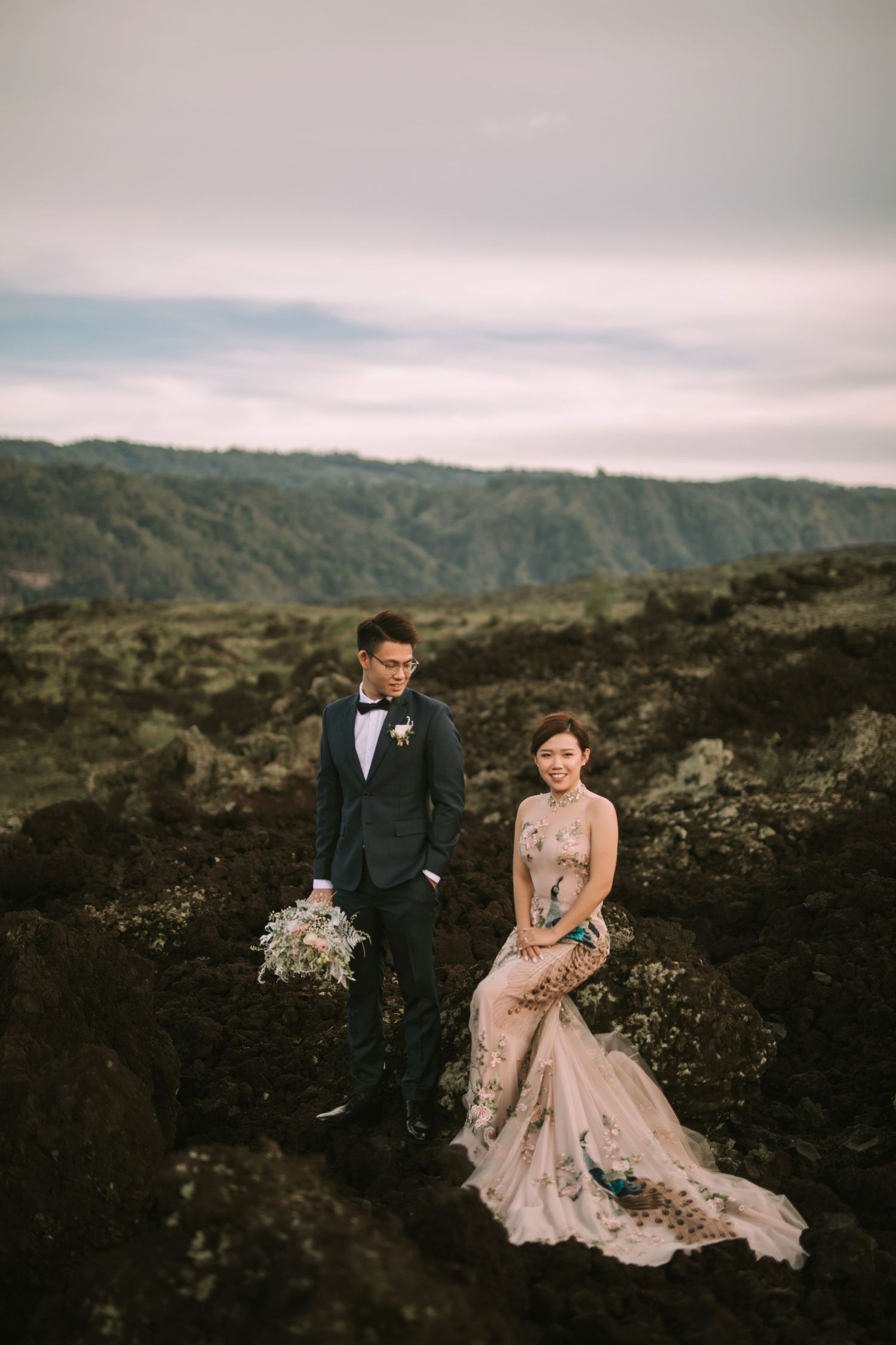 Wedding couple poses on dark volcanic rocks with cloudy mountain backdrop.