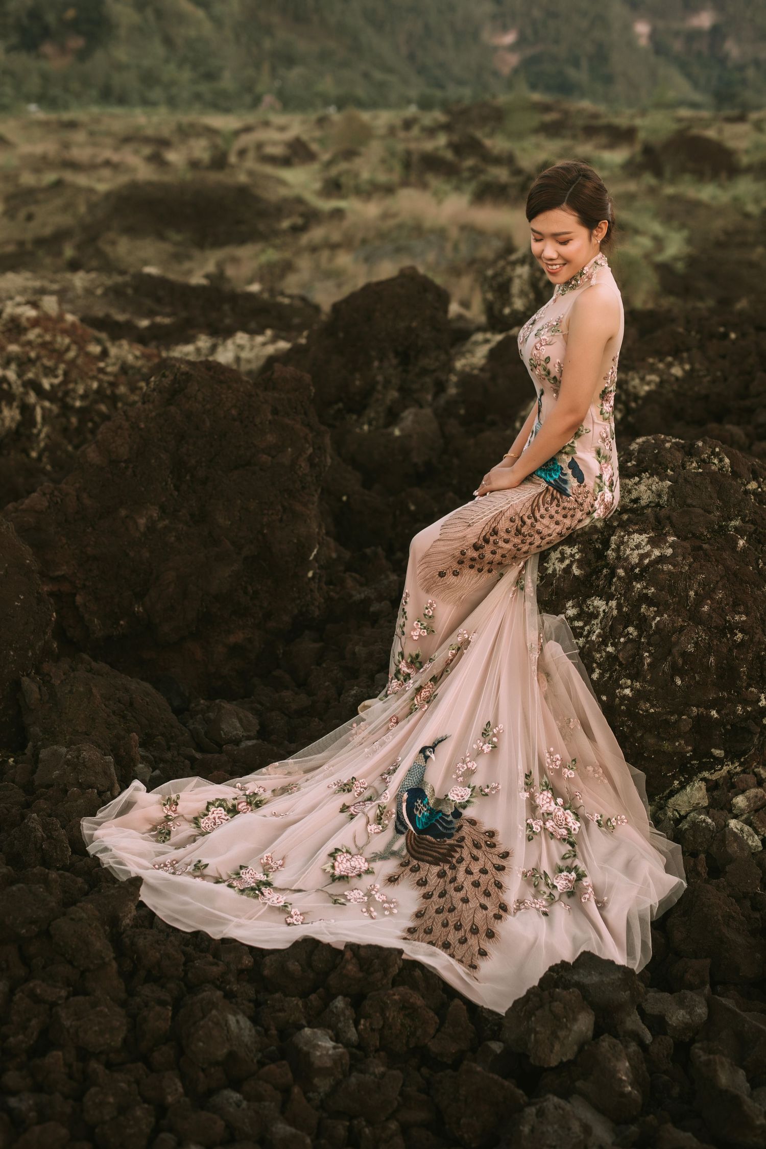 Flowing pink embellished wedding dress spreads across dark volcanic rocks in dramatic landscape.