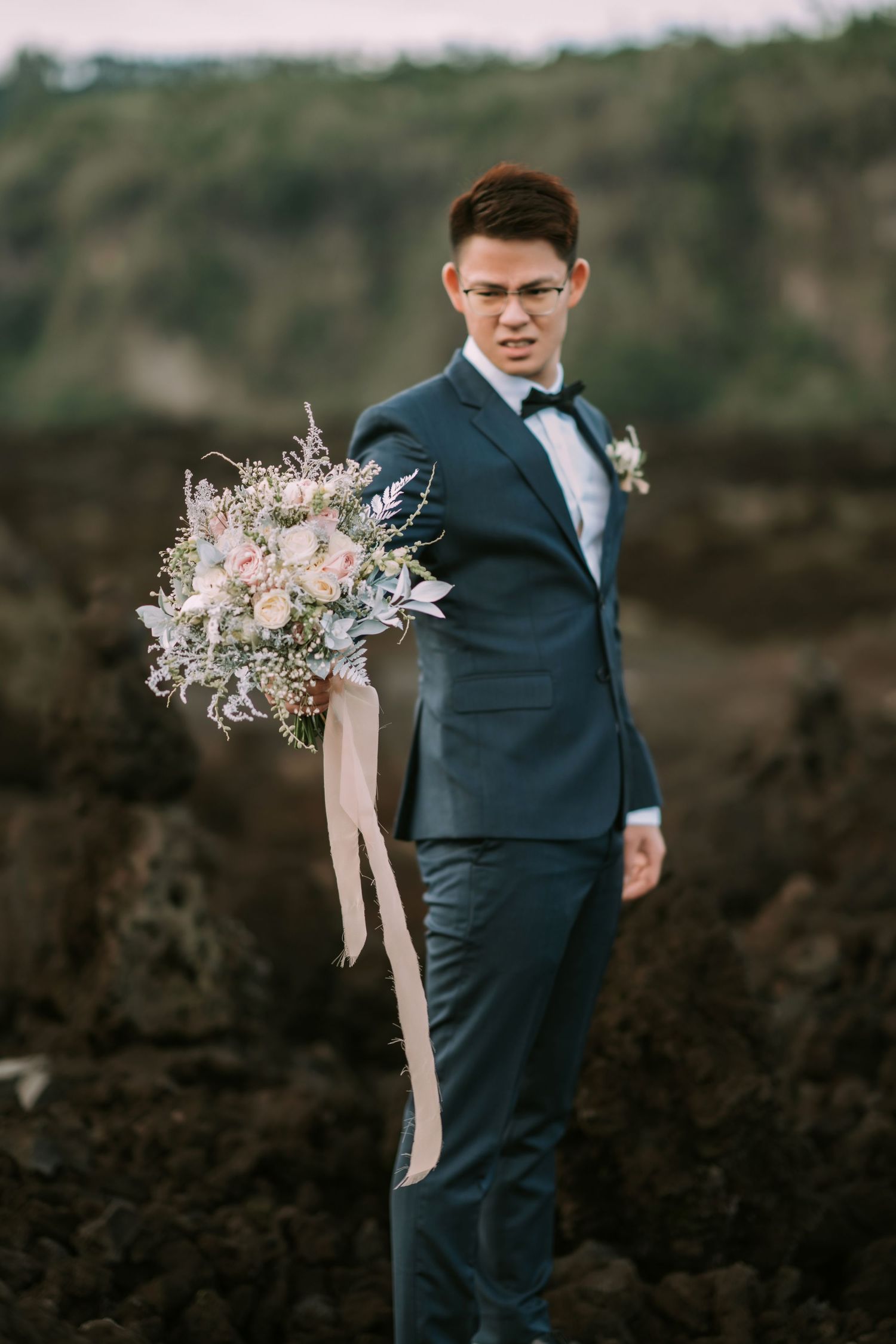 Navy suited figure holds delicate floral bouquet against volcanic landscape backdrop.