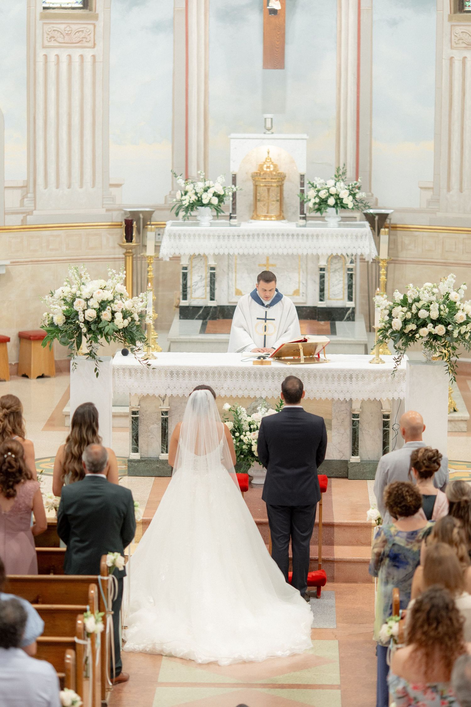 Traditional Catholic wedding ceremony at ornate altar with floral arrangements and gathered guests.
