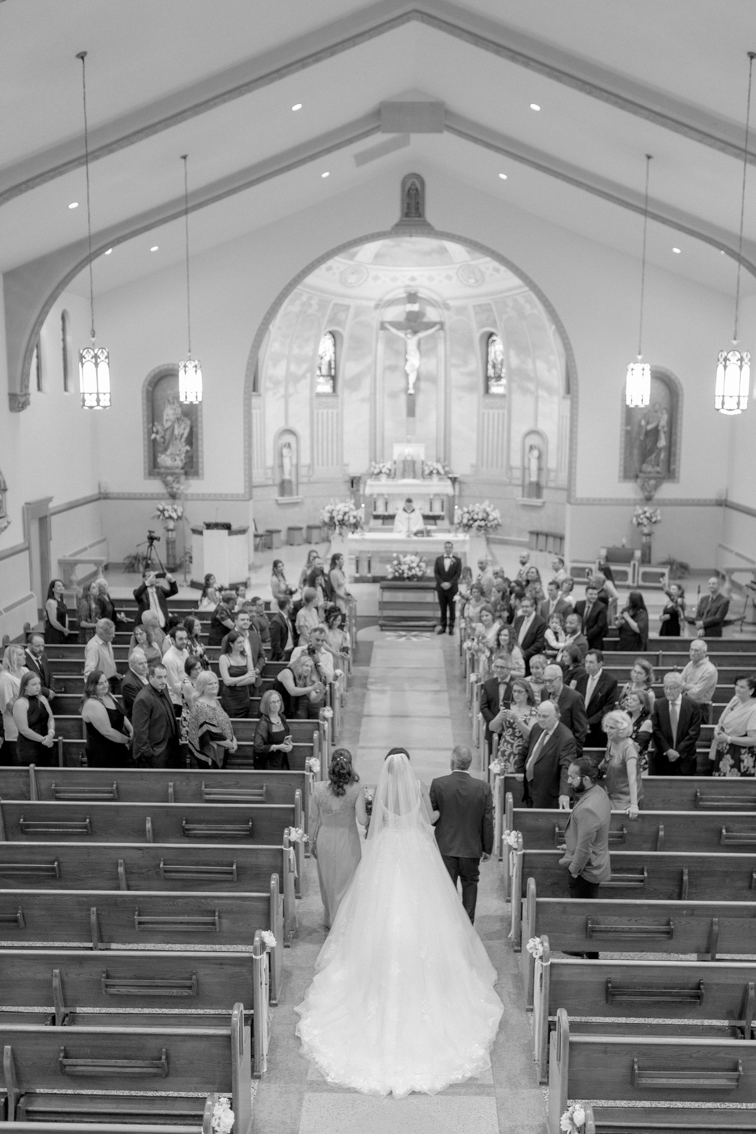 Black and white view down church aisle during traditional wedding ceremony with gathered guests.