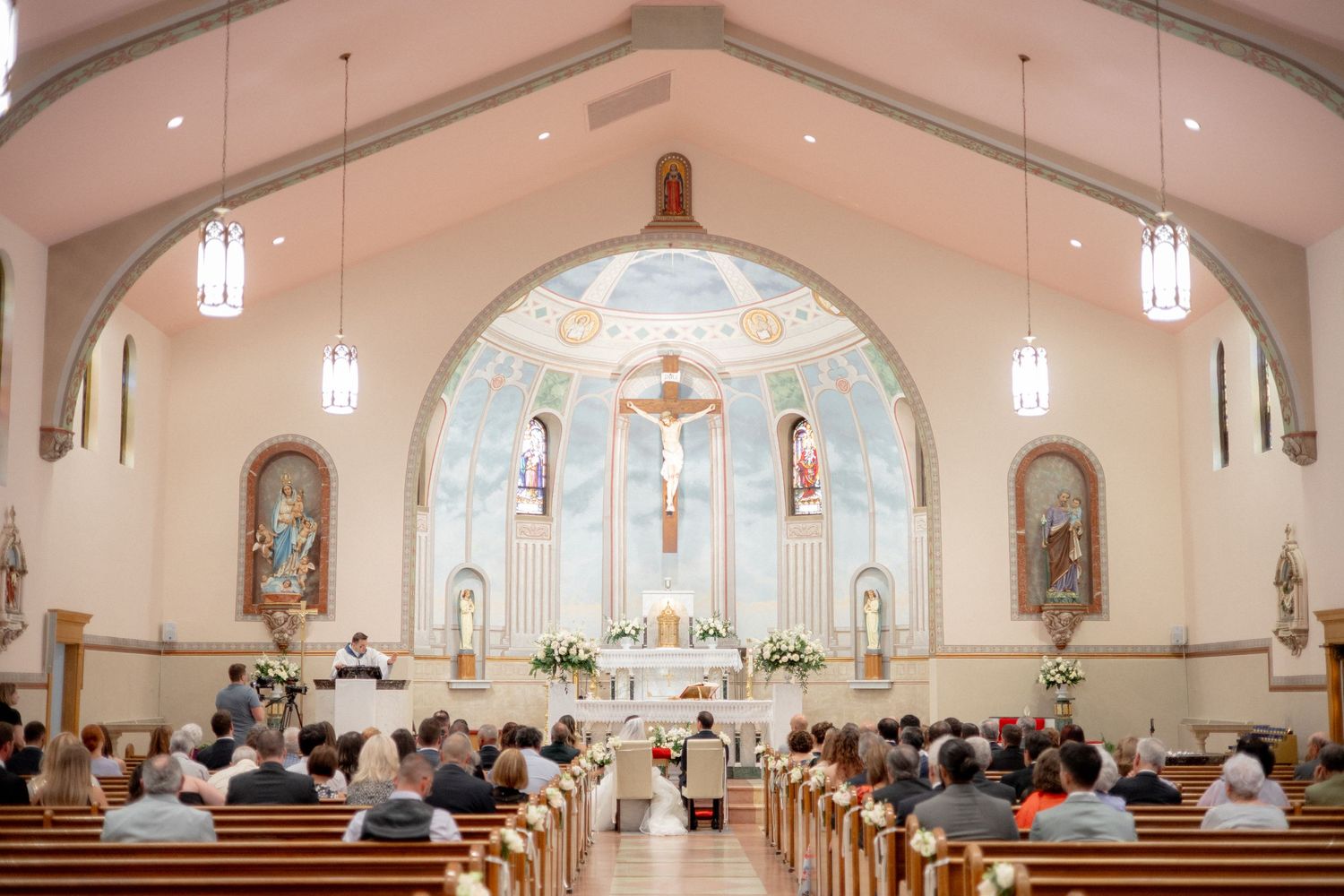 Interior of a Catholic church during a wedding ceremony with ornate altar and hanging pendant lights.