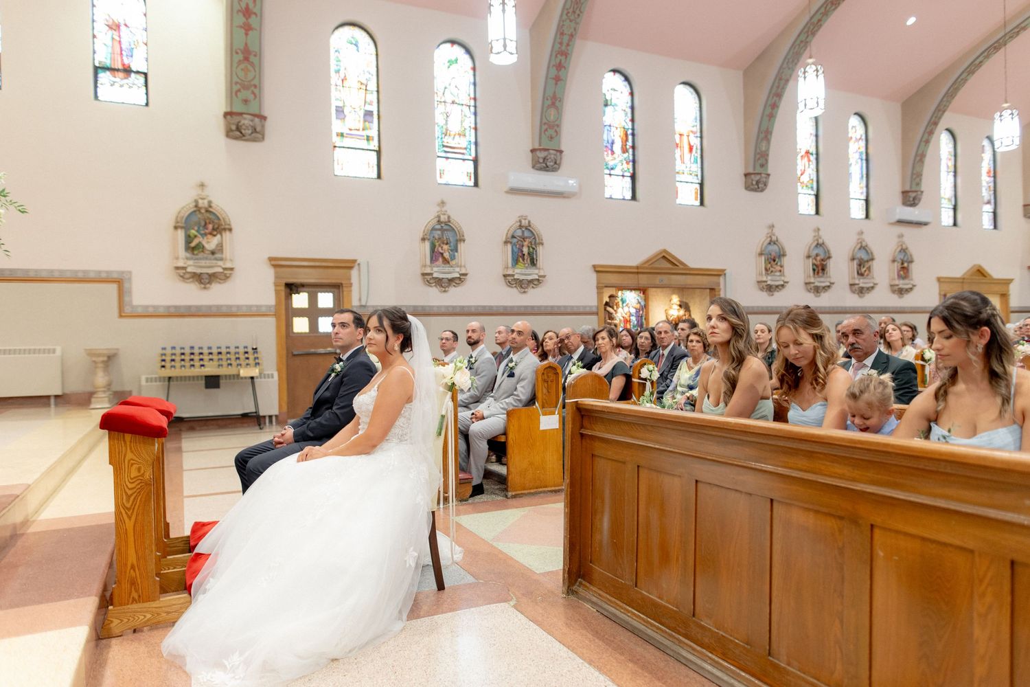 Wedding ceremony in a traditional Catholic church with stained glass windows and wooden pews filled with guests.