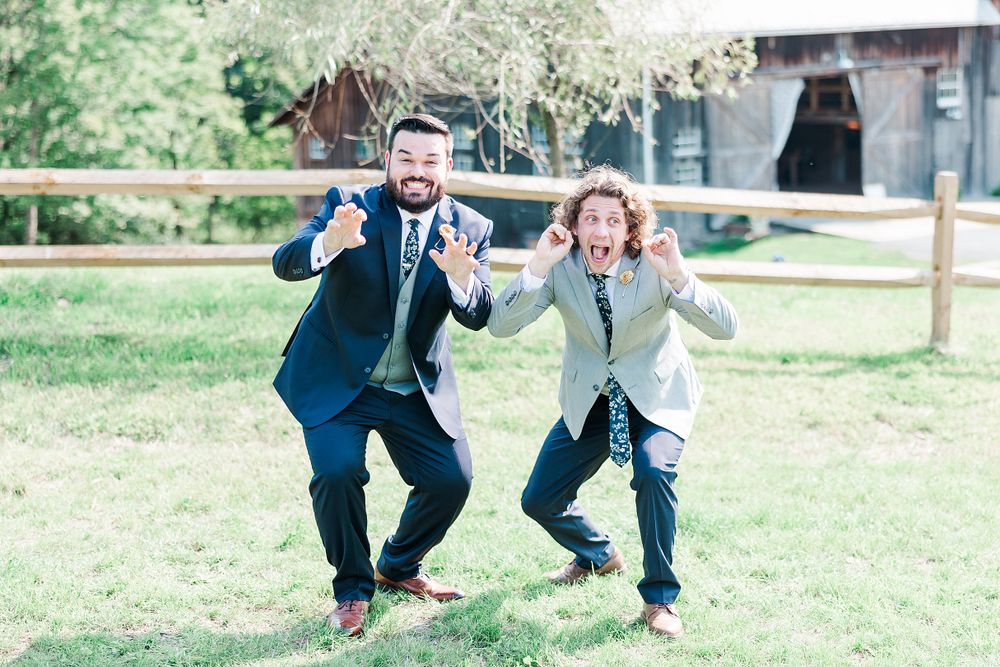 Two groomsmen in suits jump and pose energetically on the grass during wedding festivities.