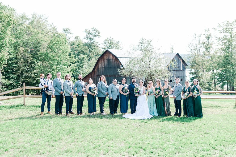 Wedding party poses in front of rustic barn with bridesmaids in green dresses and groomsmen in gray.