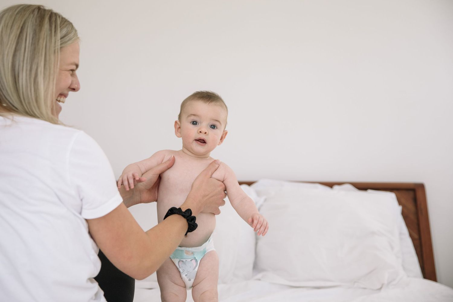 Baby in diaper sits on white bed while being held.