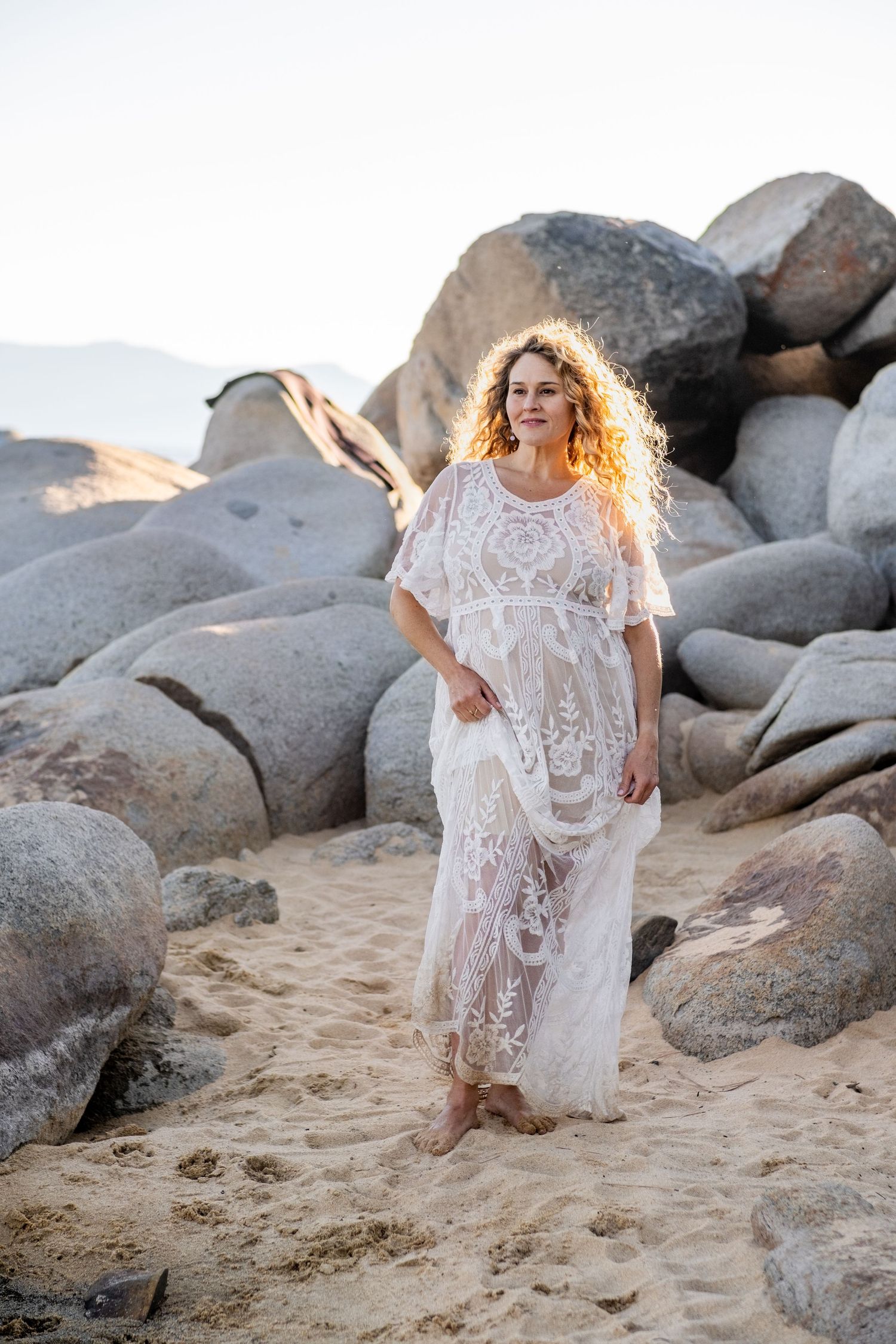 A pregnant woman wearing an elegant white lace maxi dress flows in the wind as sunlight illuminates the rocky beach setting at golden hour.