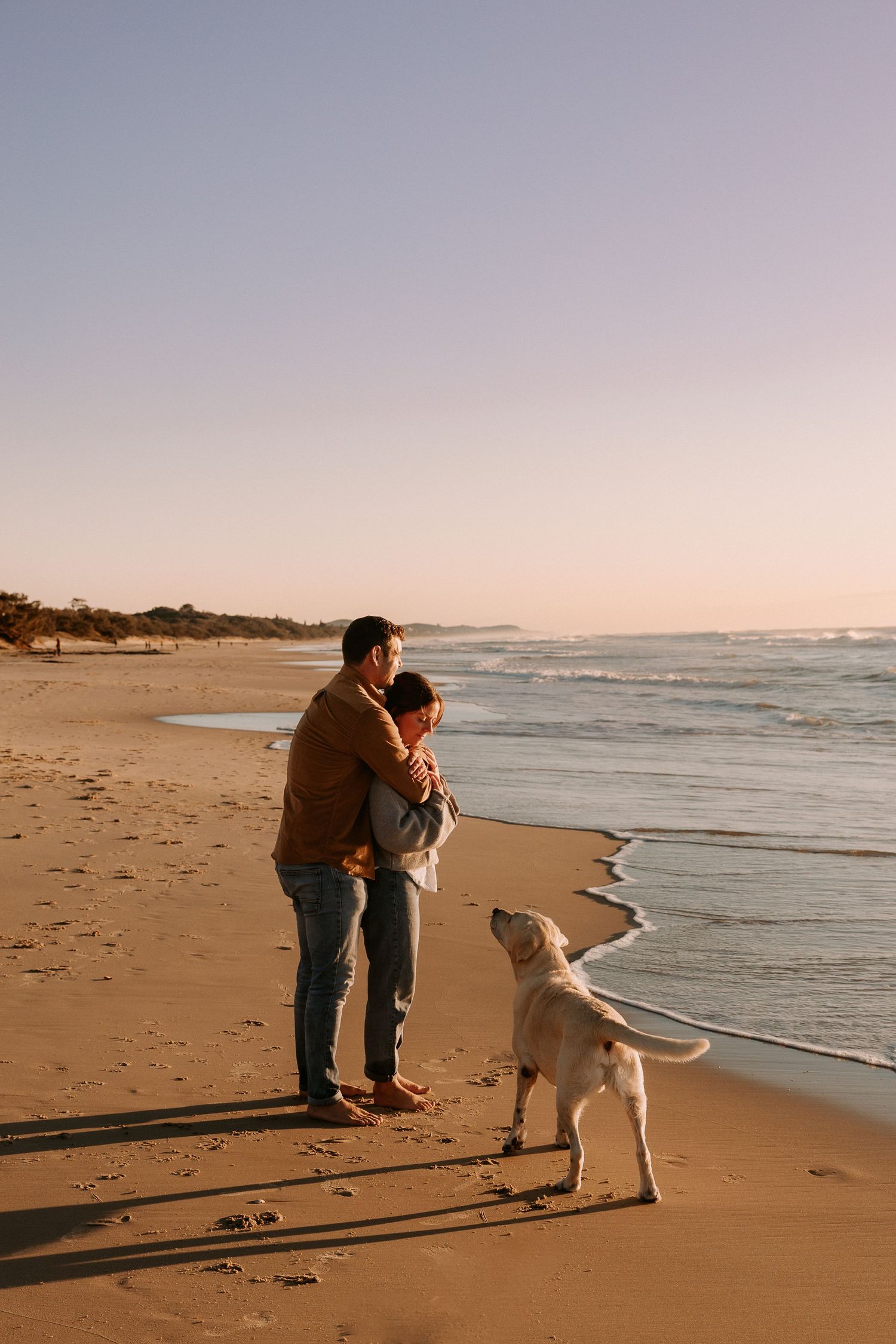 A couple and their dog walking along the shoreline at sunset.