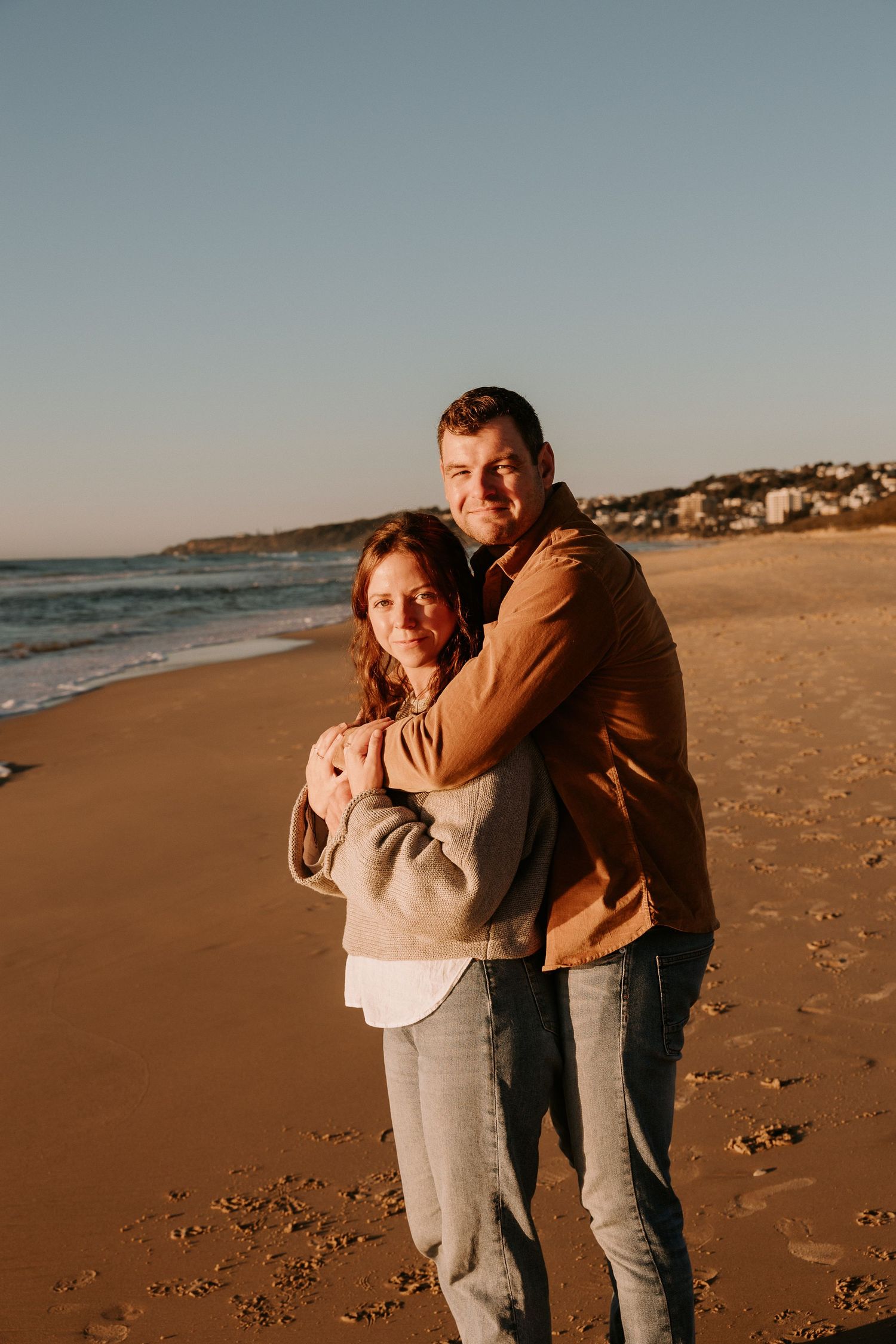 A couple embraces on a golden-lit beach during sunset.