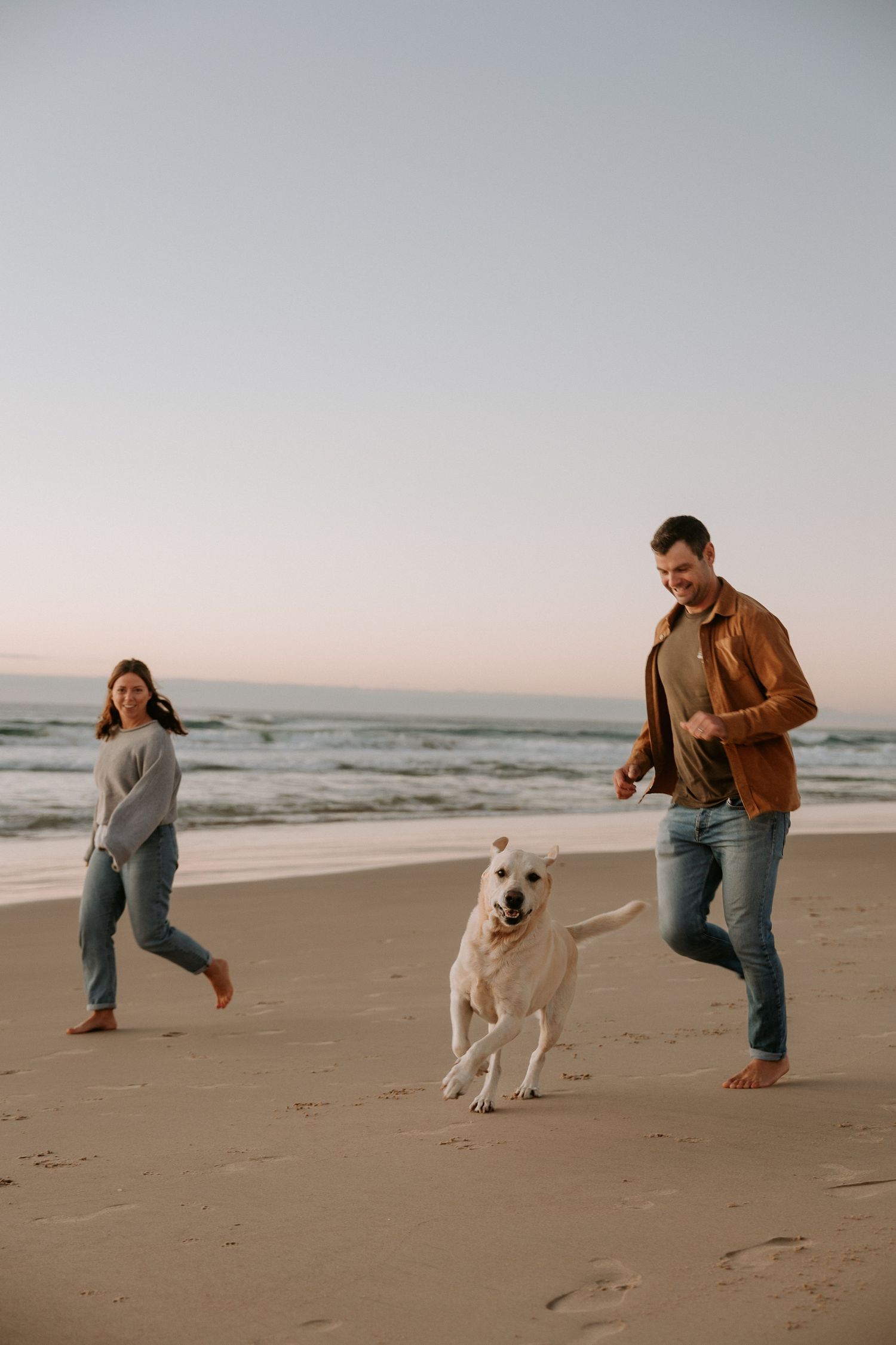 Happy couple and their energetic dog running together on sandy beach at sunset.