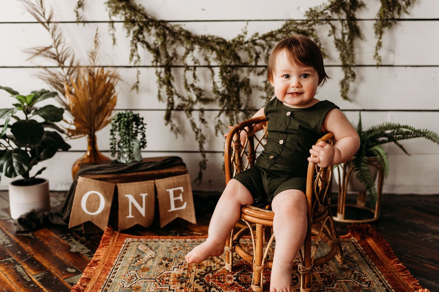 Baby in green romper sitting on wooden chair with rustic decor and 'ONE' blocks.