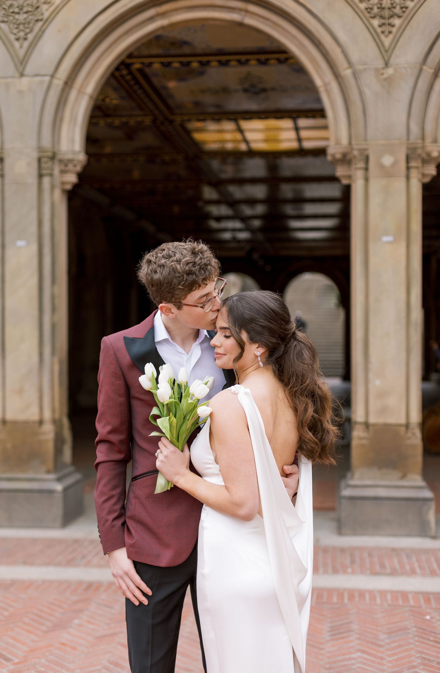 A couple embraces beneath ornate archways holding white tulips during their wedding.