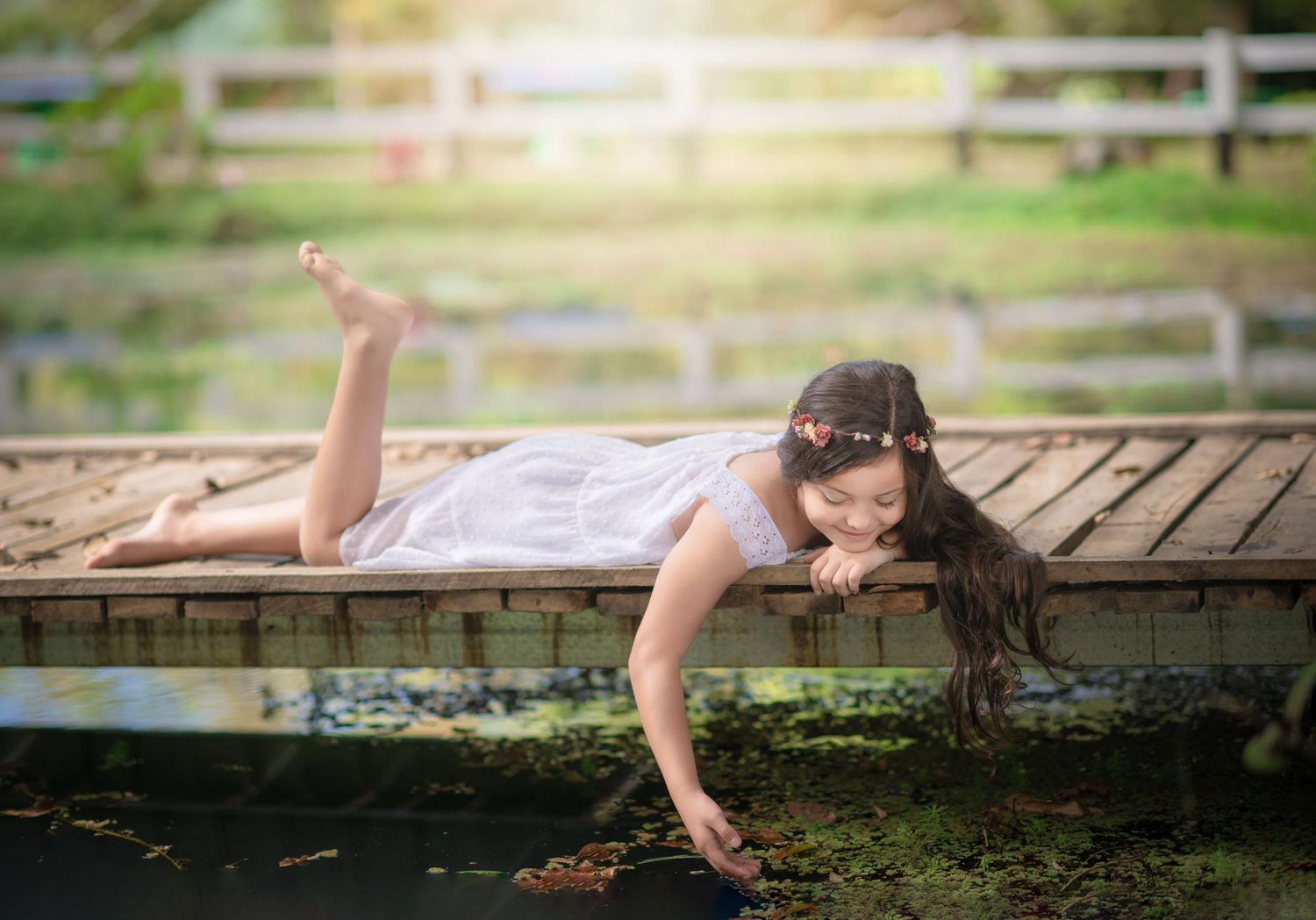 Dreamy portrait of someone lying on wooden dock over pond in white dress.