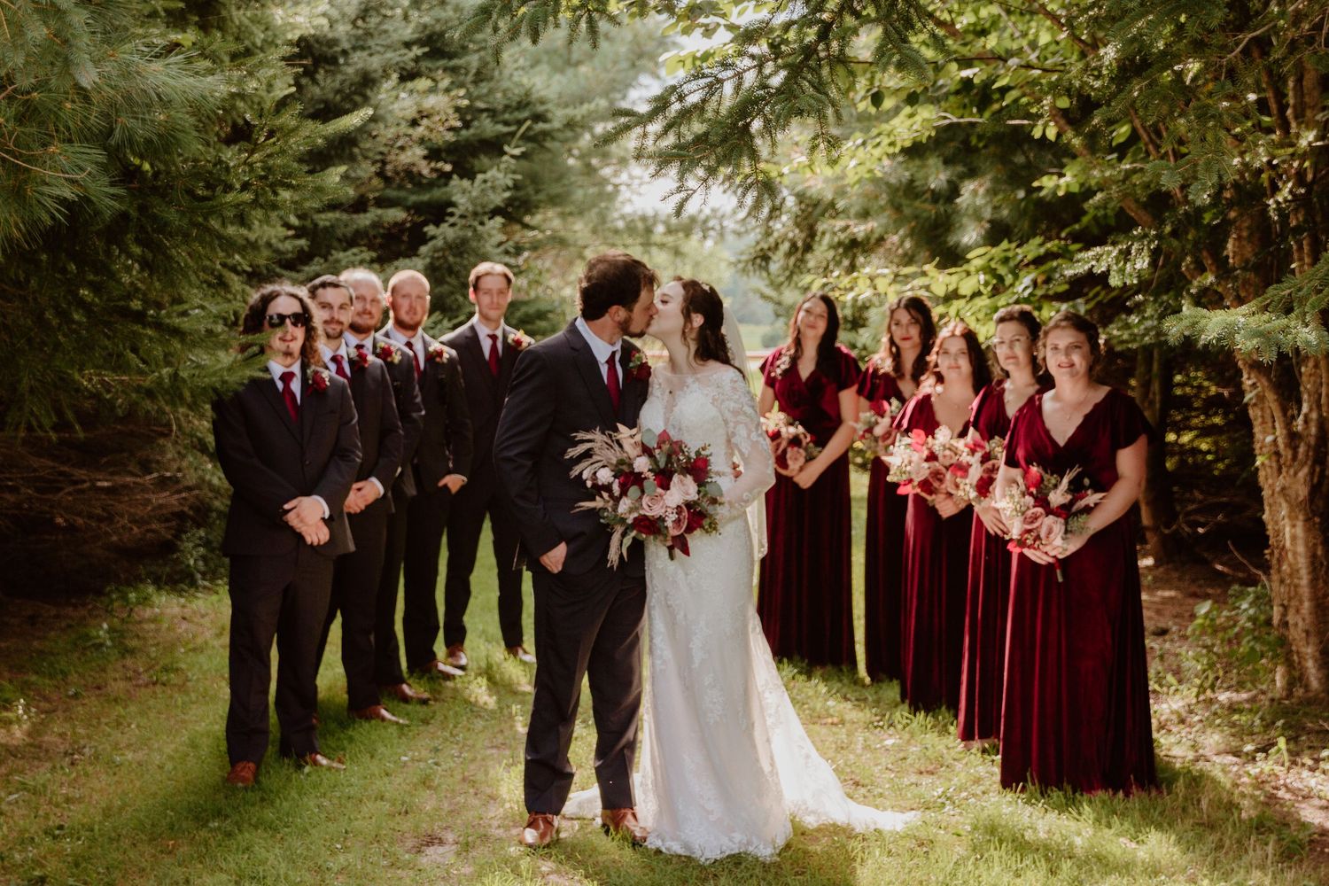 Wedding party lines up in burgundy dresses and black suits among evergreen trees.