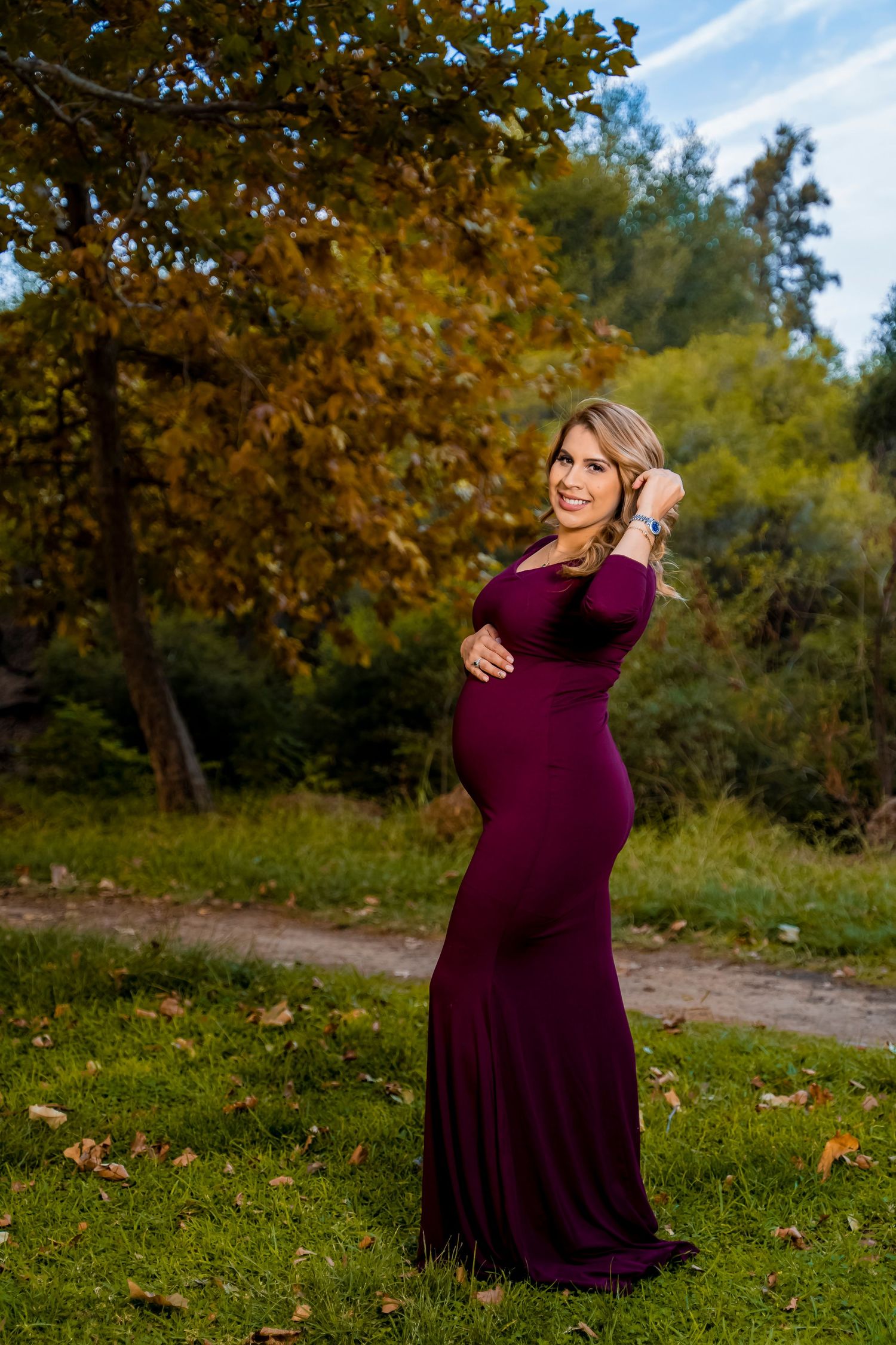 A burgundy floor-length maternity gown worn outdoors during autumn with golden foliage in the background.