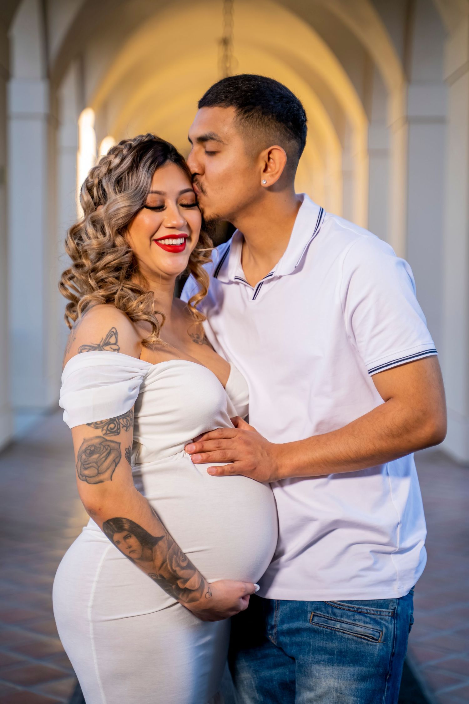 An expectant couple shares a tender moment in an arched hallway.