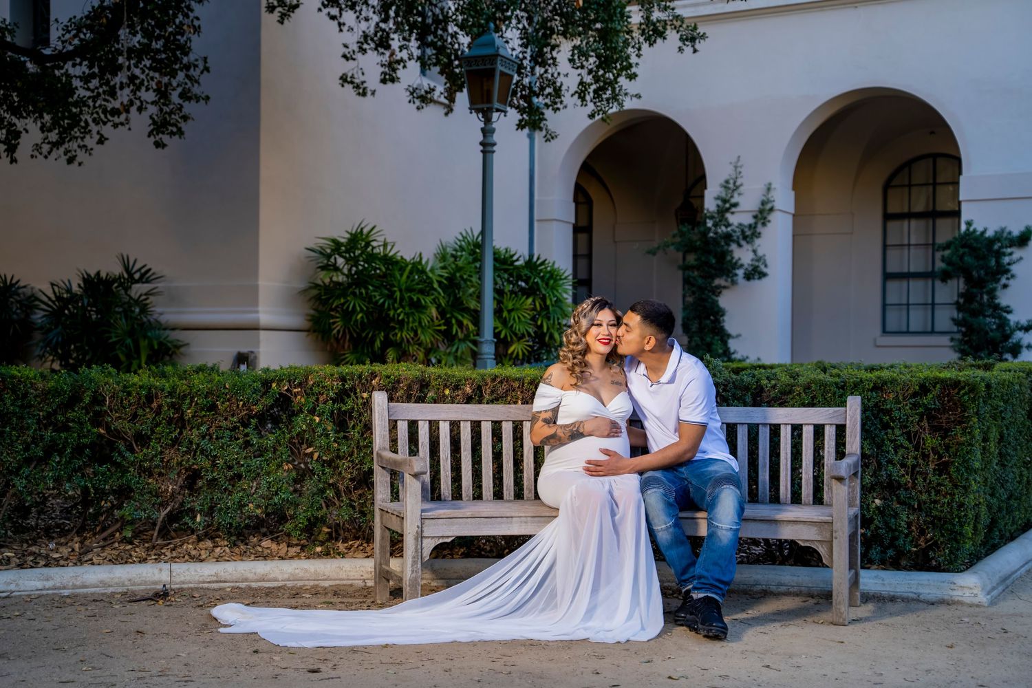 A couple shares an intimate moment on a wooden bench in front of an elegant white building.