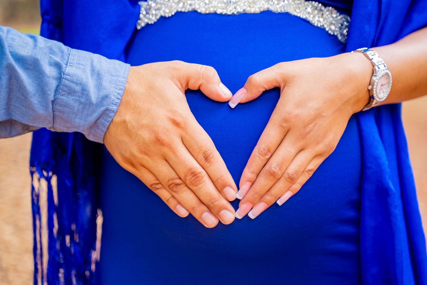 Hands forming heart shape over pregnant belly in blue dress.