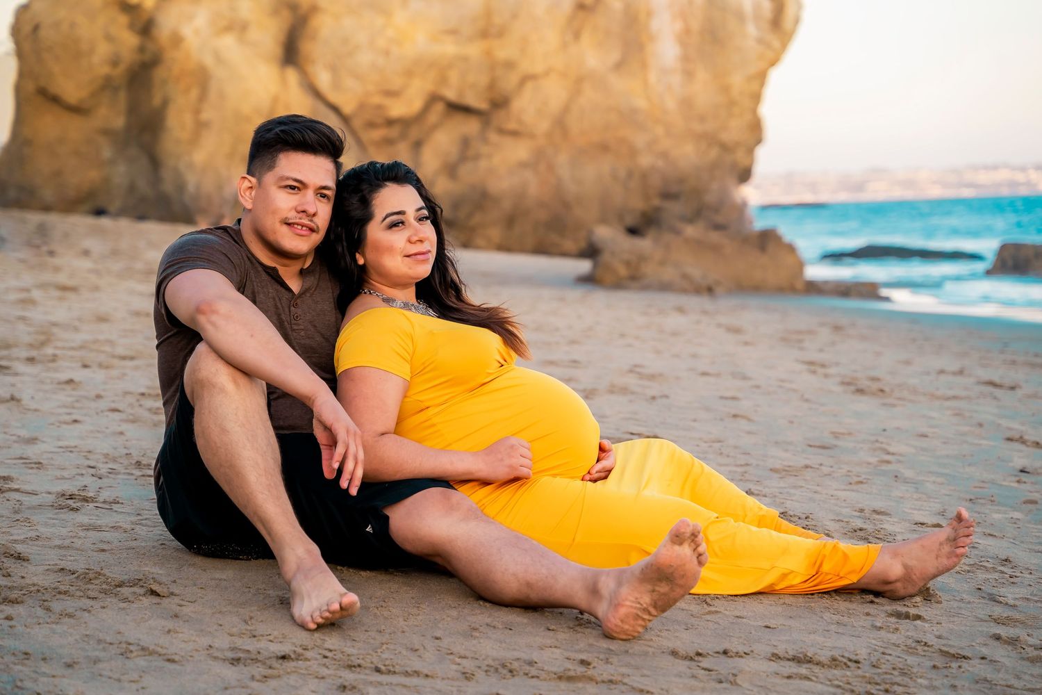 A relaxed maternity beach portrait with rocky cliffs and ocean waves in the background.