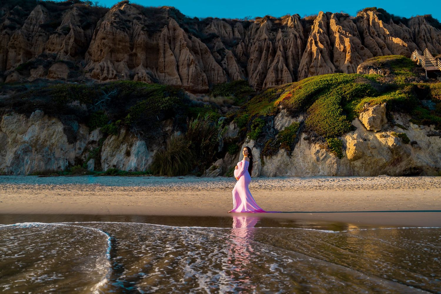 A flowing pink dress catches the light as waves lap at a rocky beach during sunset.