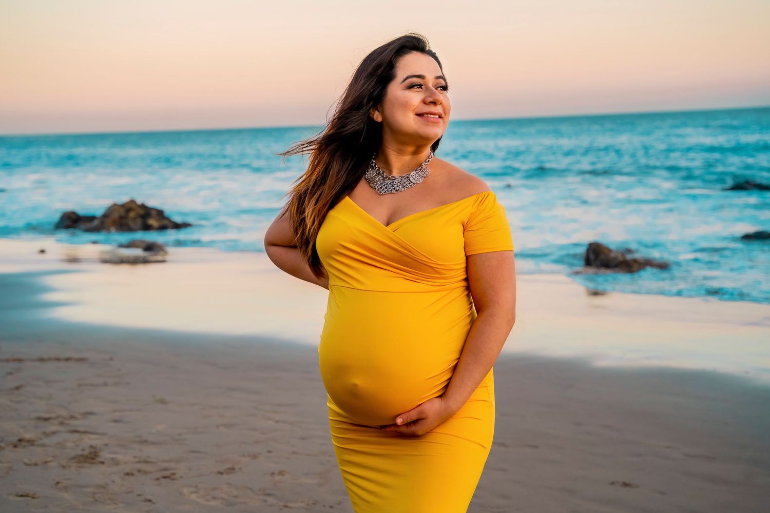 A yellow dress maternity photo shoot at sunset on a beach with crashing waves and rocks.