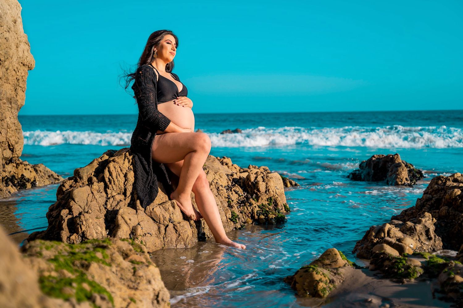 Maternity photo shoot on rocky beach with turquoise ocean waves in background.