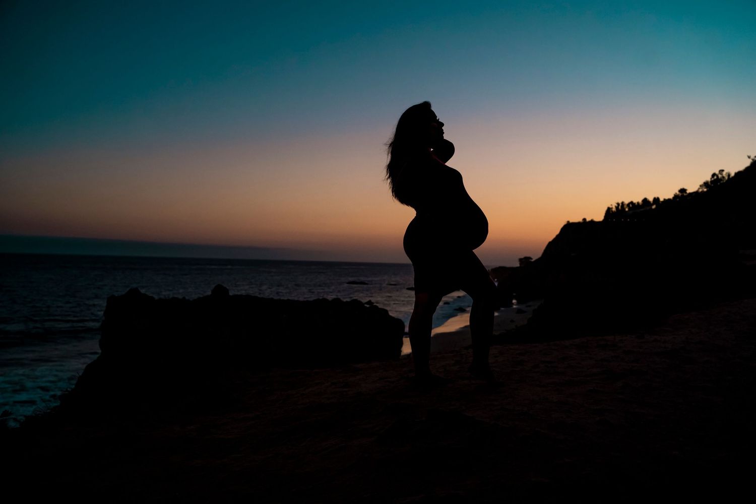 A silhouette against sunset sky shows a dramatic maternity pose on coastal cliffs.