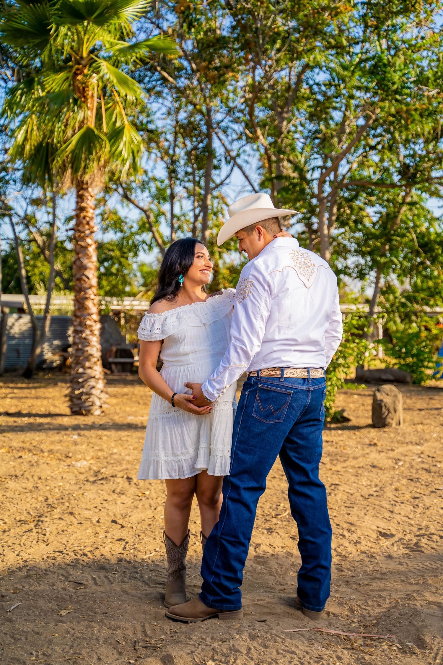 Romantic maternity moment under palm trees in rustic outdoor setting.