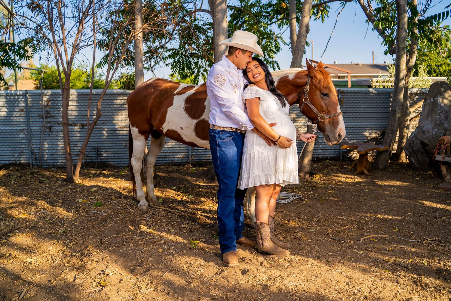 Expecting couple poses with paint horse in rustic ranch setting with palm trees.