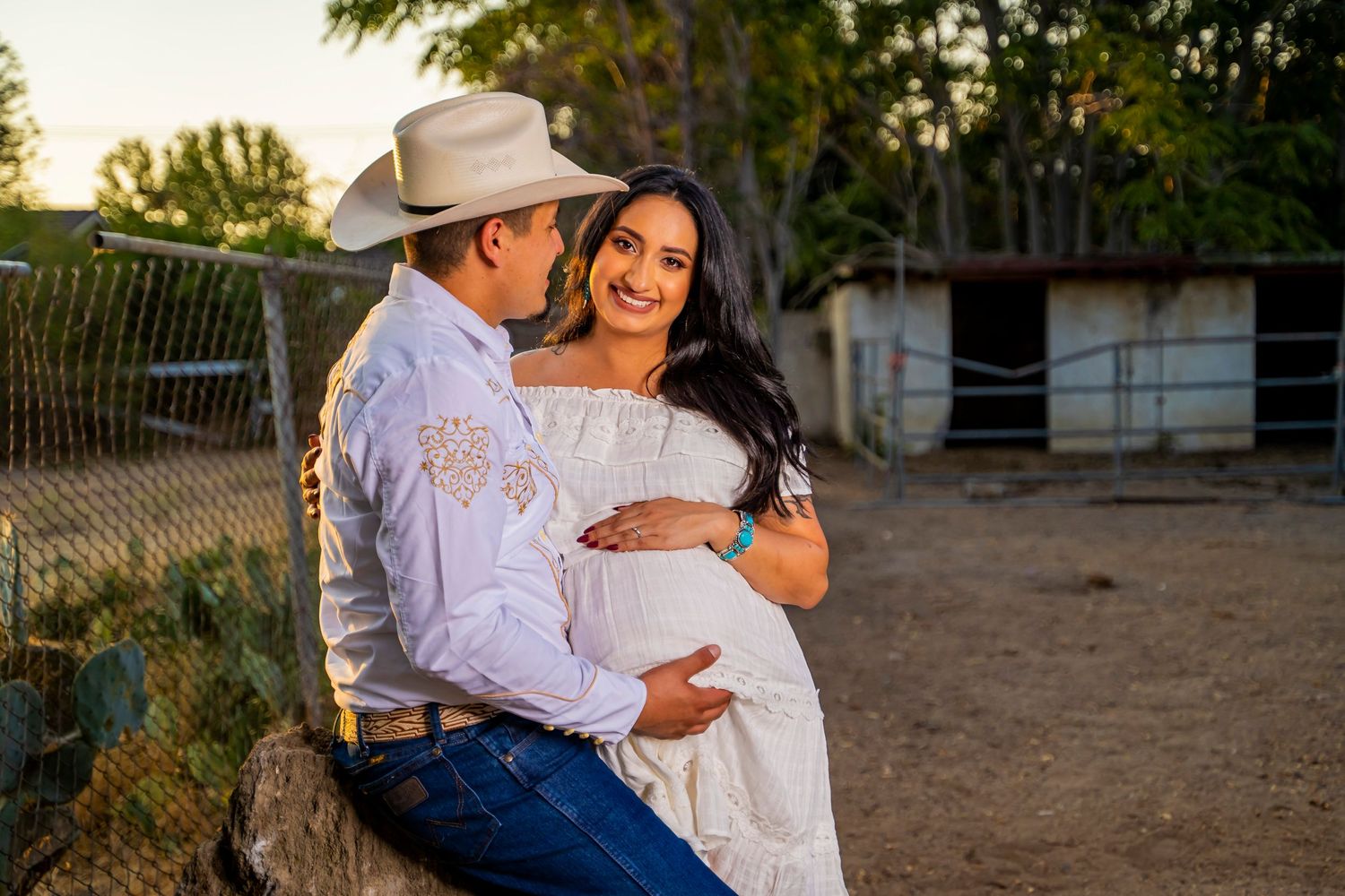 A couple embraces tenderly against a rustic fence during a sunset maternity photoshoot.