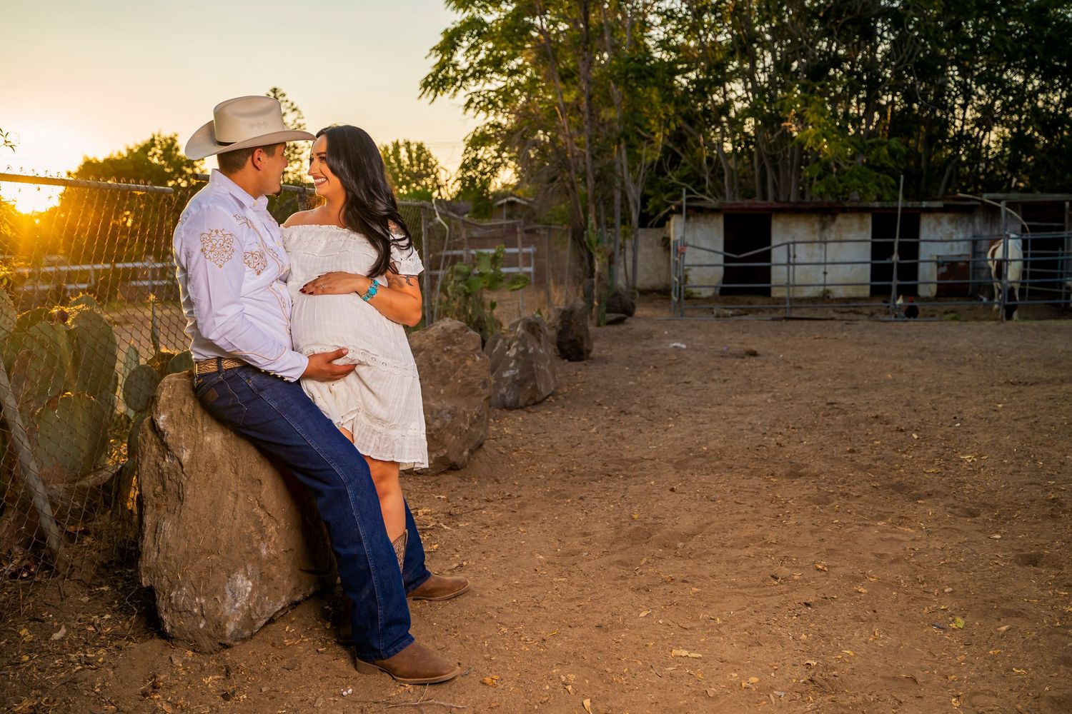 A couple shares a romantic sunset moment on rustic rocks near a barn in a countryside setting.