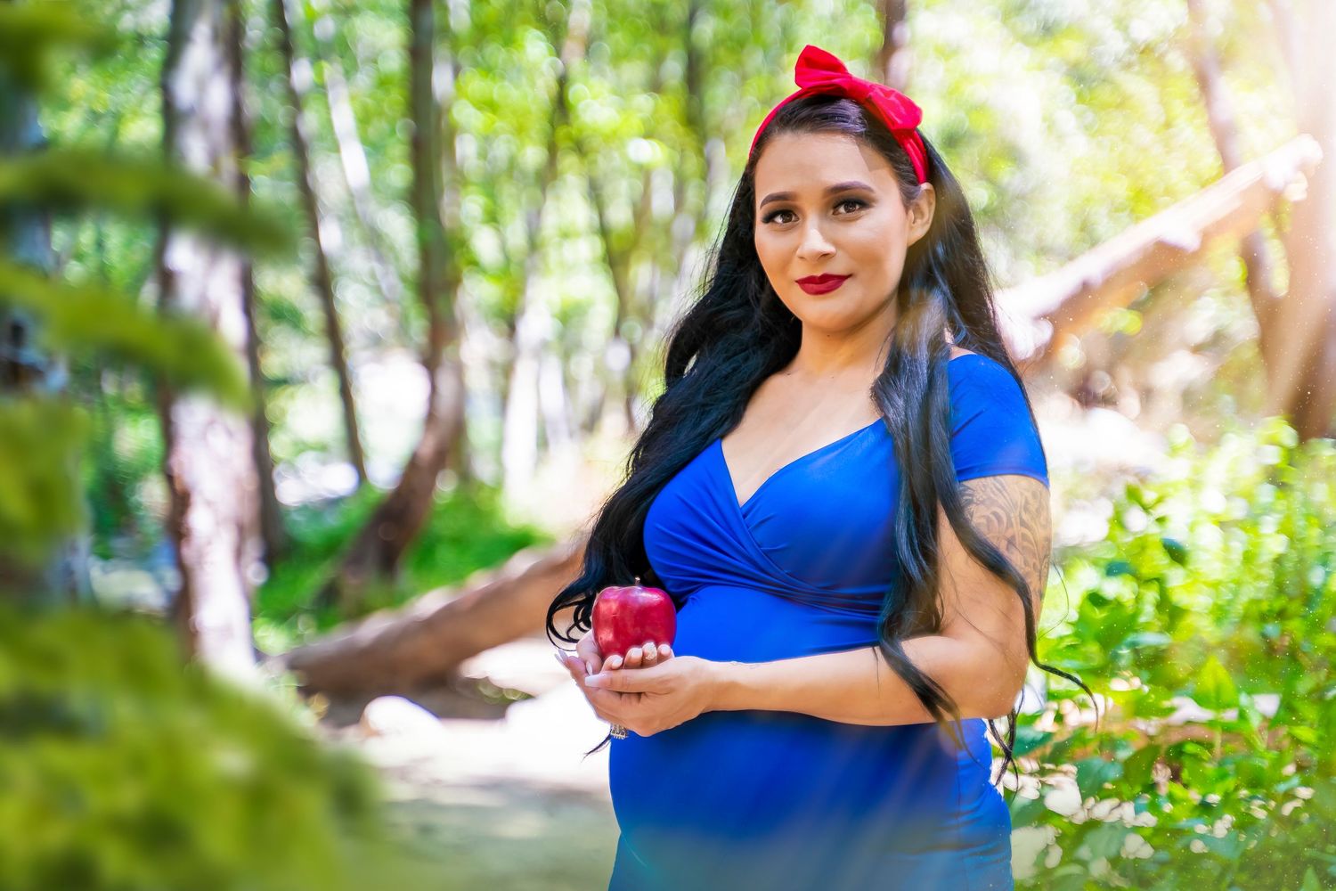 Expectant mother in blue dress poses outdoors holding small red box.