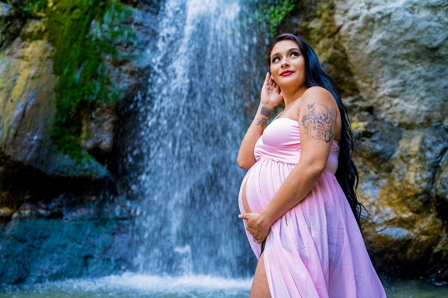 Maternity photo of expectant mom in pink dress standing by a scenic waterfall.