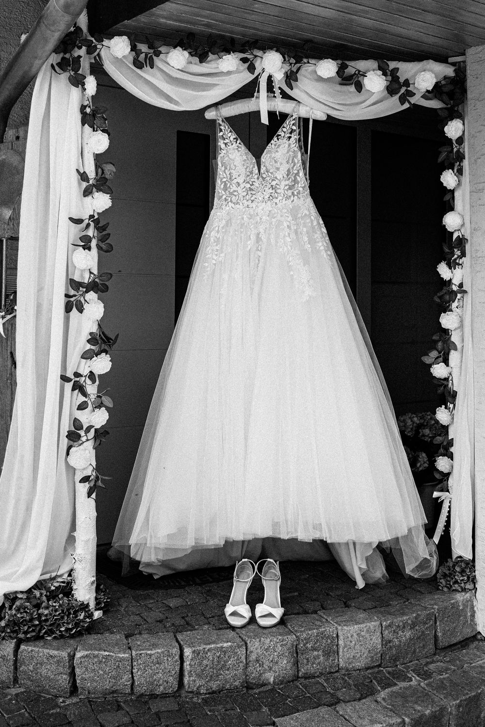 Wedding dress hanging in decorated archway with bridal shoes beneath.