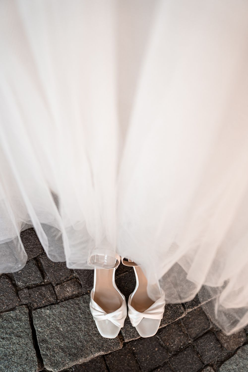 White bridal shoes placed beneath flowing tulle wedding dress on stone pavement.