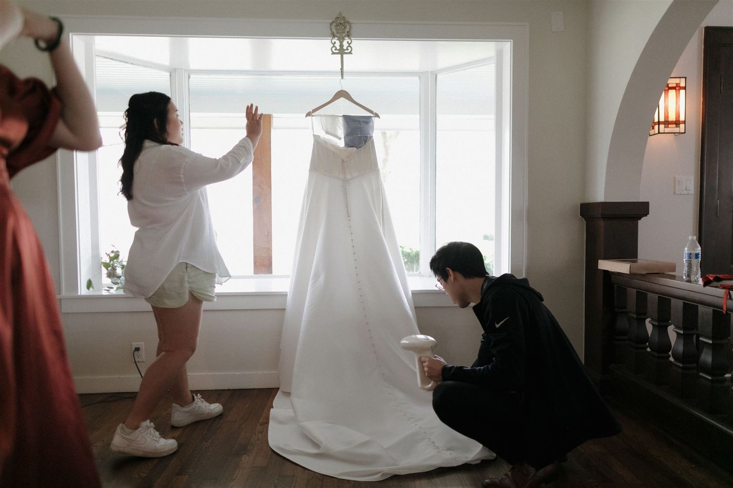 Wedding preparation scene showing people arranging a white wedding dress hanging by a window.