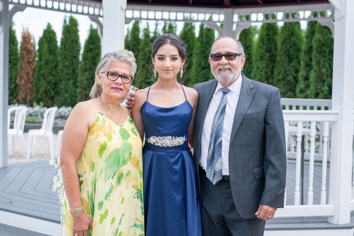 Three people pose together at outdoor venue with white gazebo and evergreen trees.