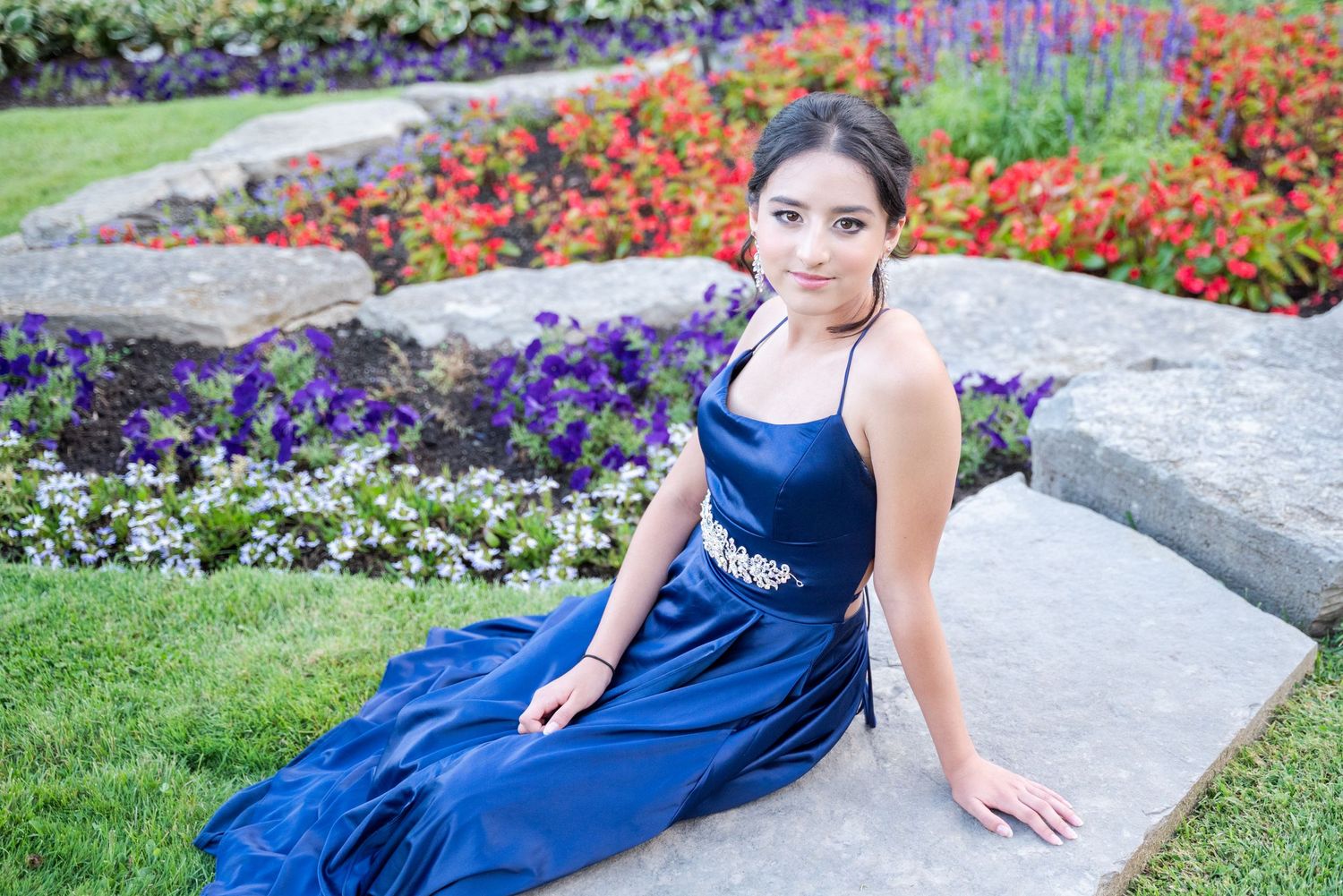 Elegant navy blue formal dress worn on stone steps amidst colorful garden flowers.
