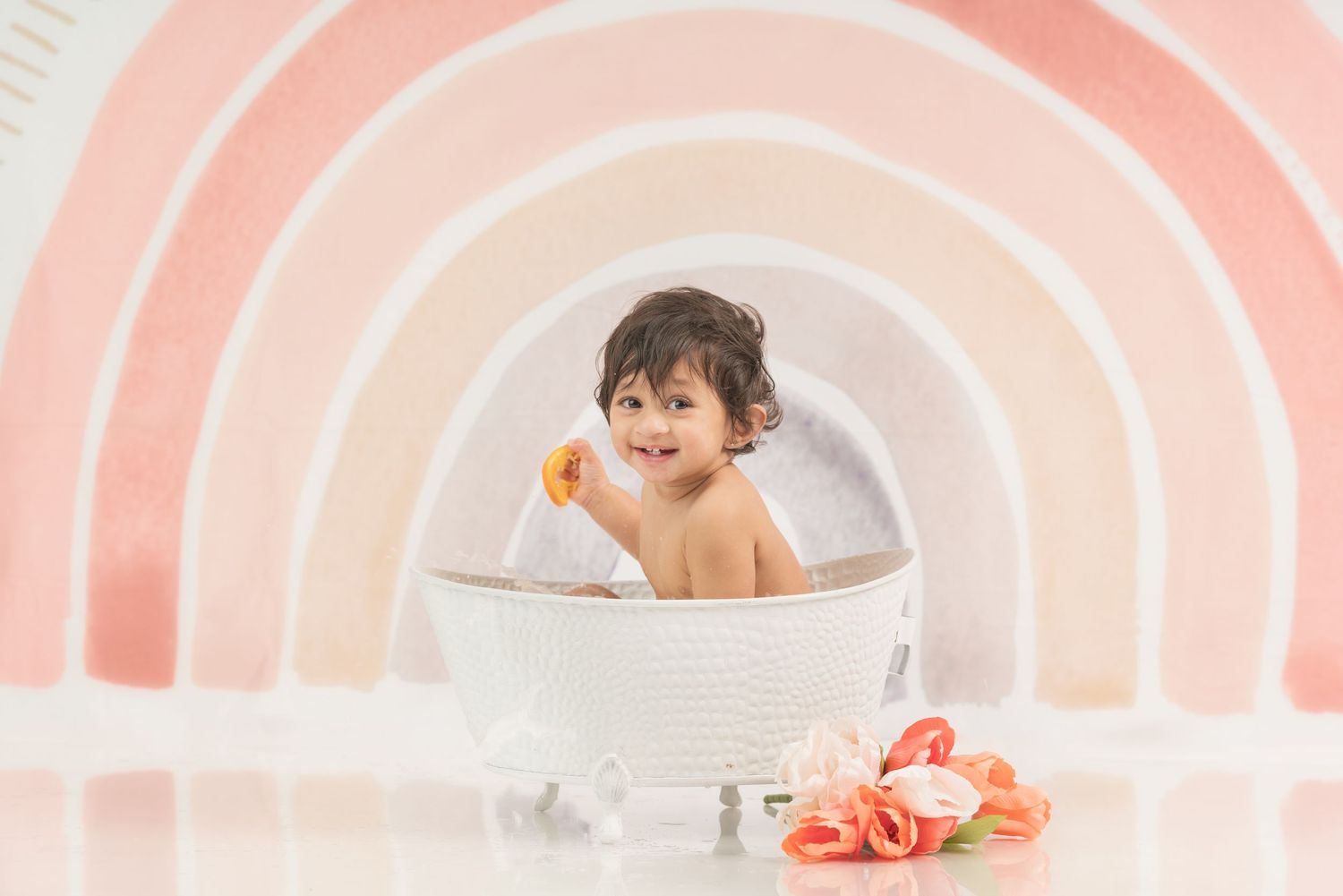 Baby in white ceramic tub with oranges against pink rainbow backdrop during photo session.