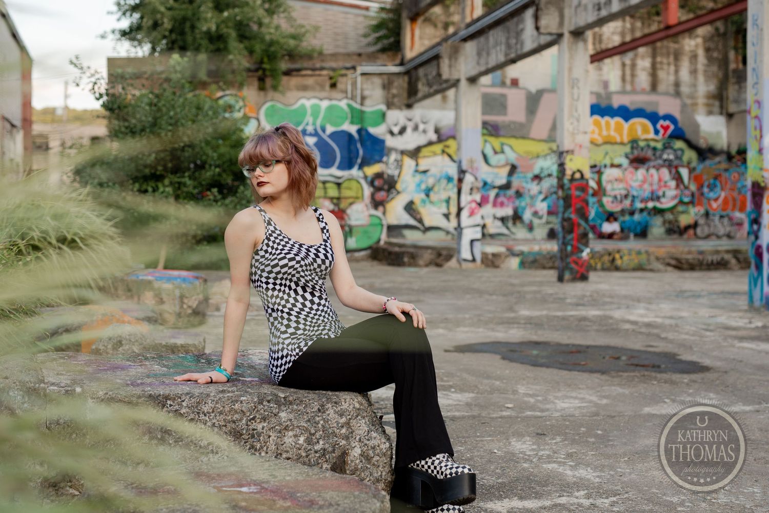 Urban fashion shoot with model seated on concrete ledge surrounded by graffiti art and foliage.