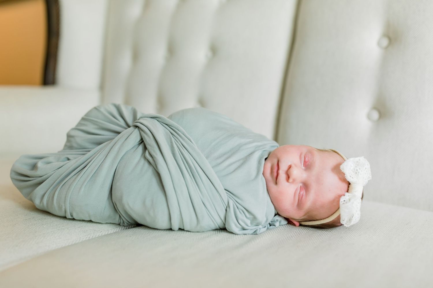 Sleeping newborn in sage green swaddle wrap resting on white tufted couch.