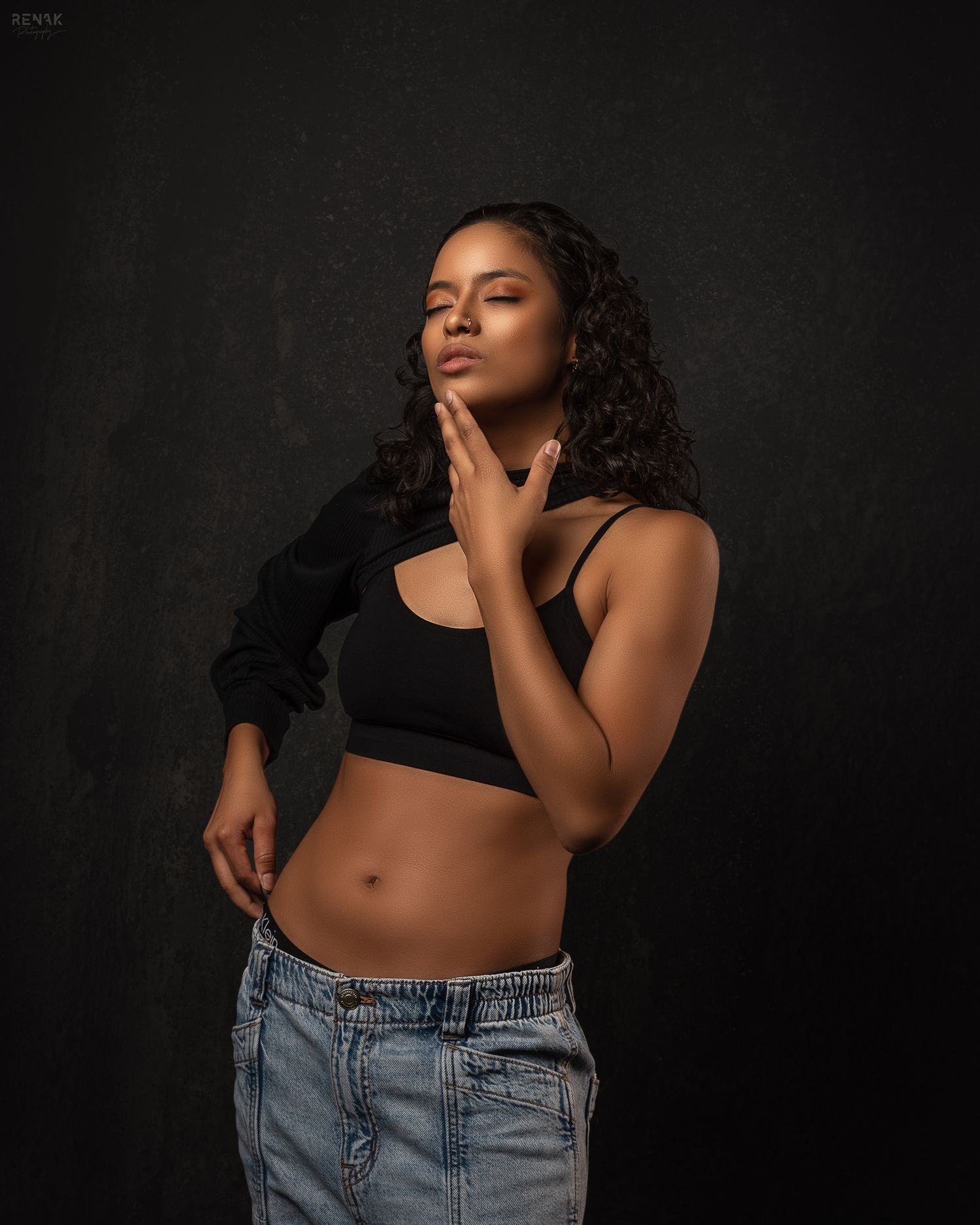 Studio portrait of someone posing in a black crop top and denim against dark background.