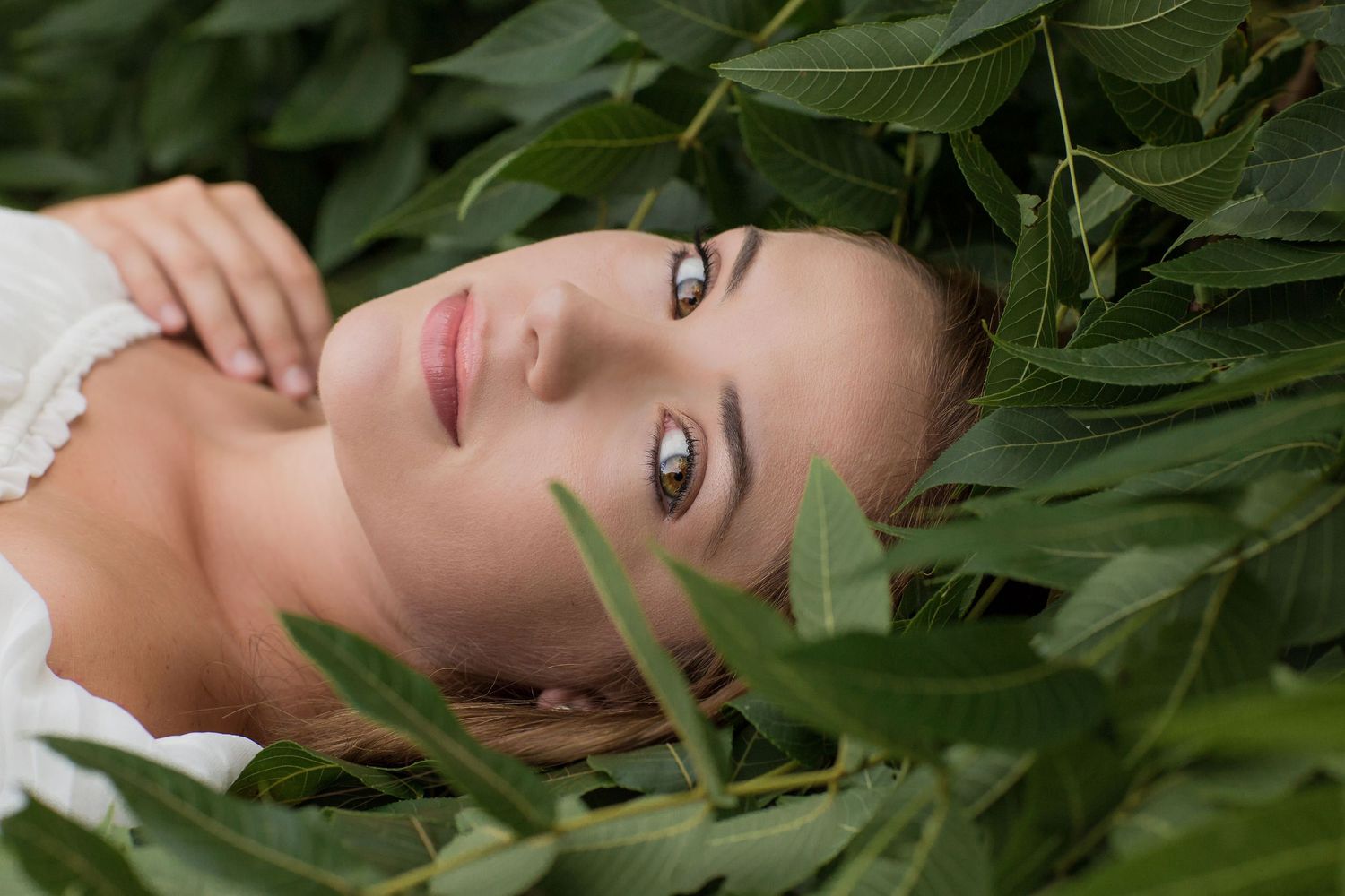 Young woman with natural makeup lying among lush green leaves in an outdoor nature portrait.