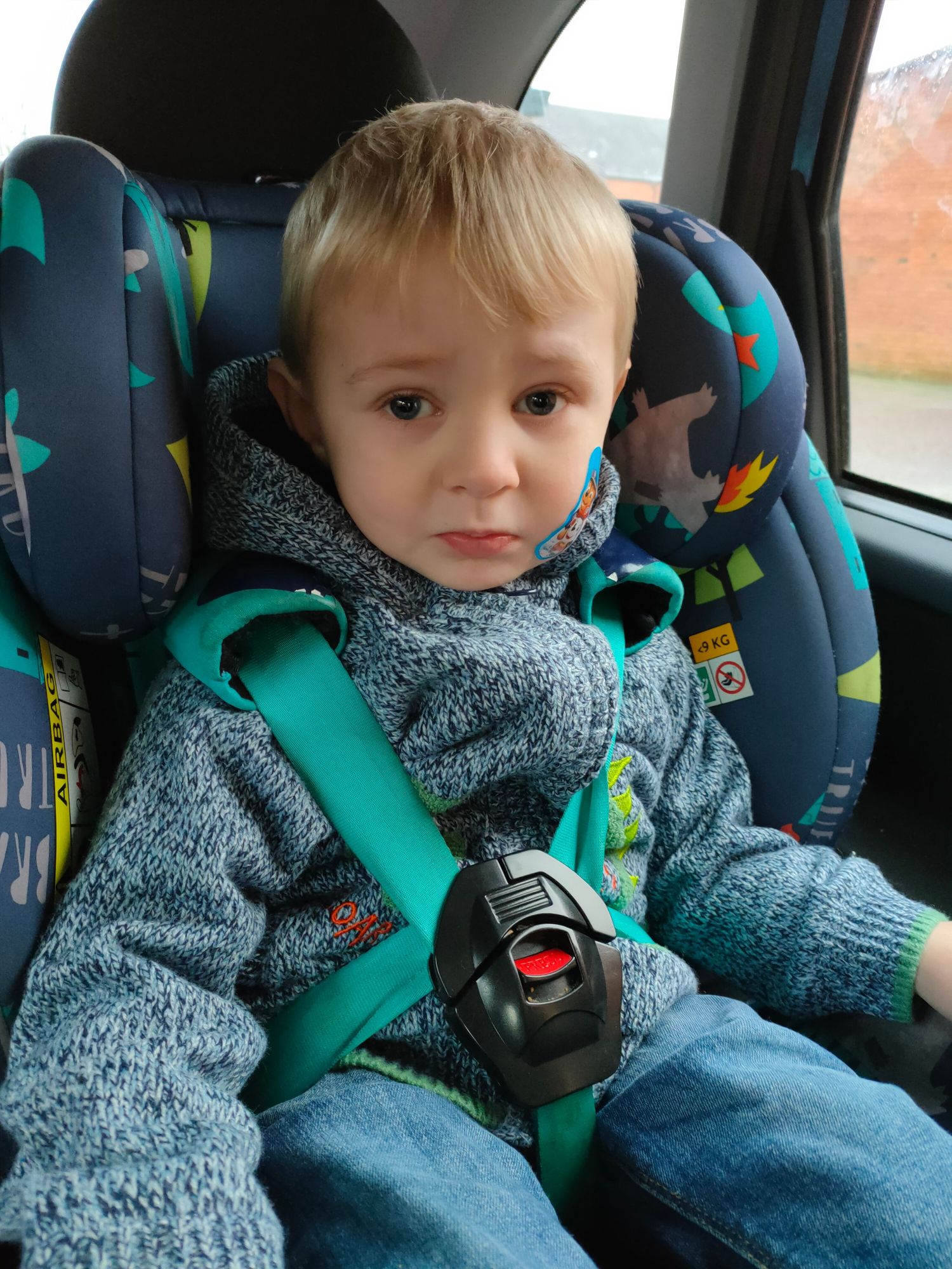 Child in gray fleece jacket secured in car seat with teal safety straps.
